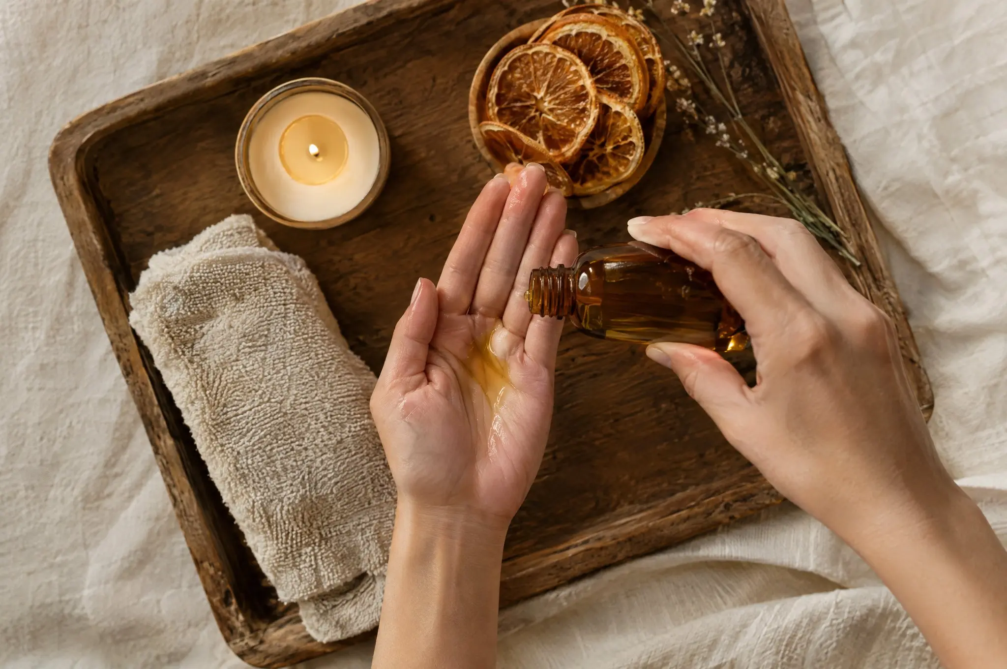 Woman pouring oil into hand, lit candle and dried oranges on wooden tray